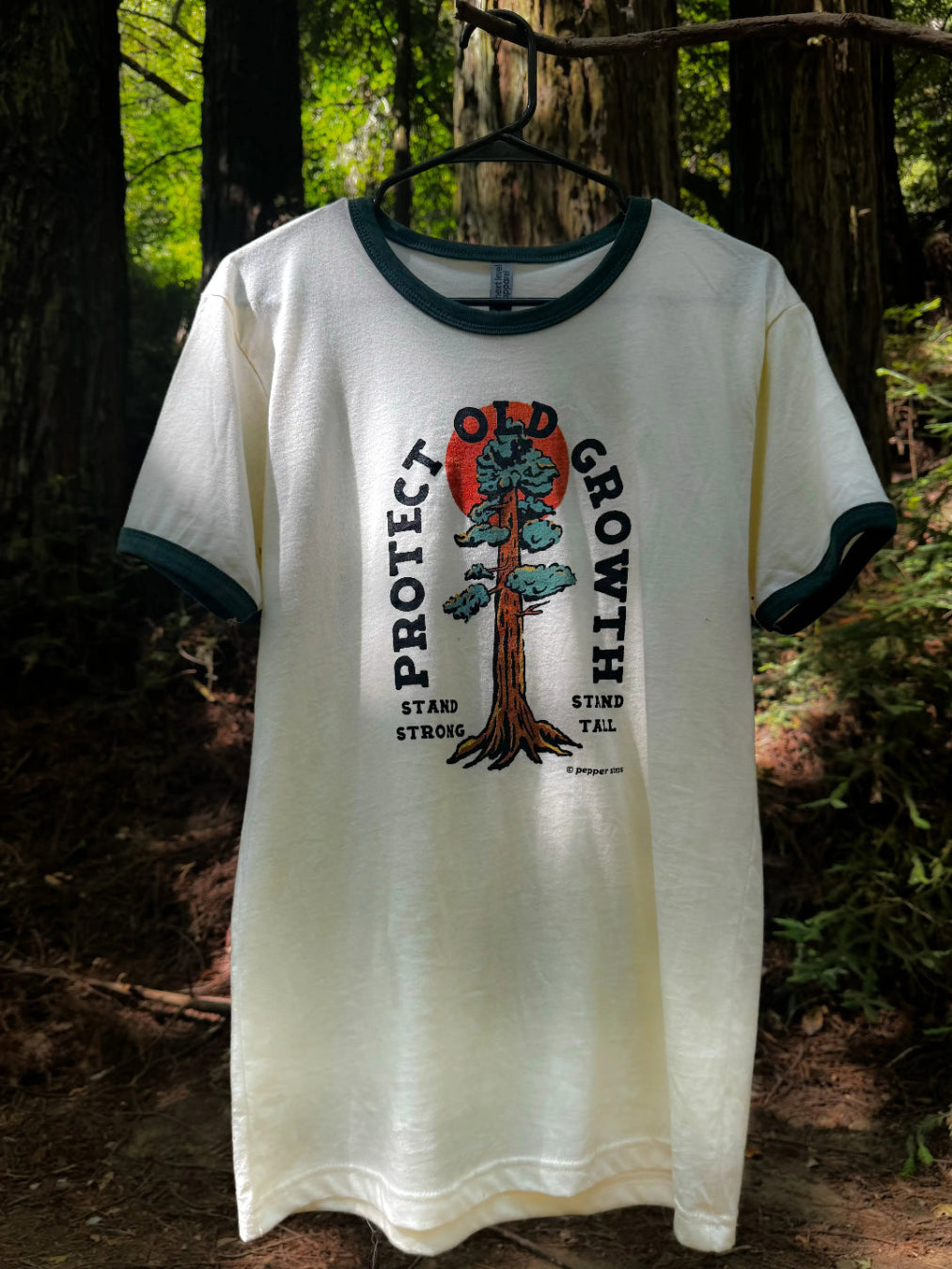 White t-shirt with a tree graphic and text on a hanger against a forest background
